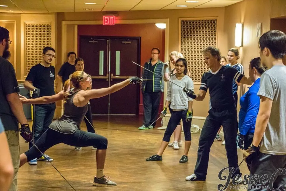 Jacqueline, surrounded by a group of people all holding rapiers, teaching a combat class