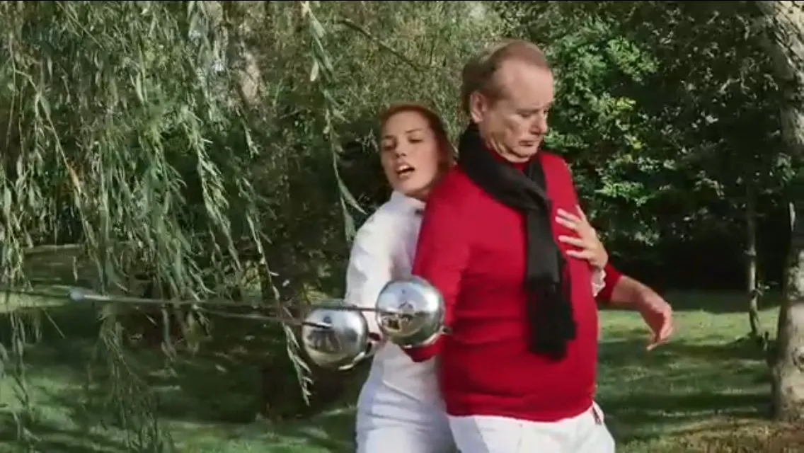 Jaqueline standing behind a man in a red shirt, both holding rapiers. 
