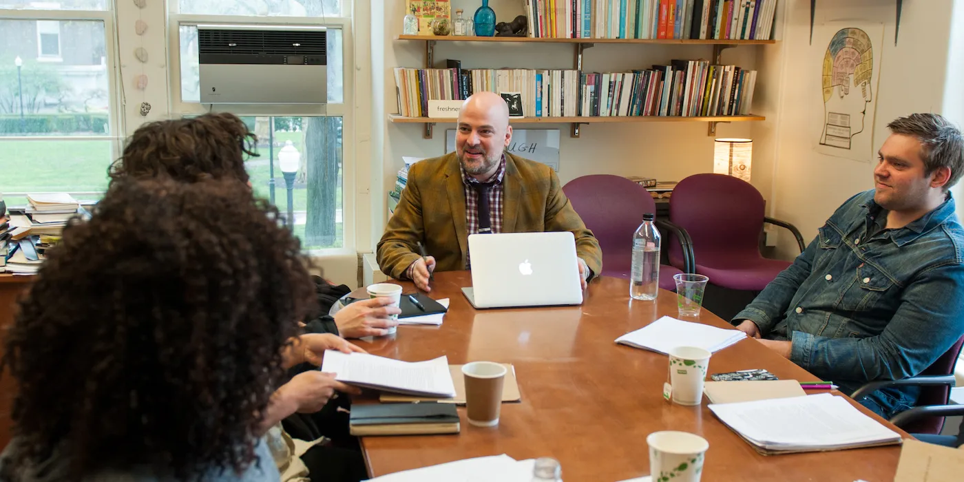 A professor sits at a table with a group of students