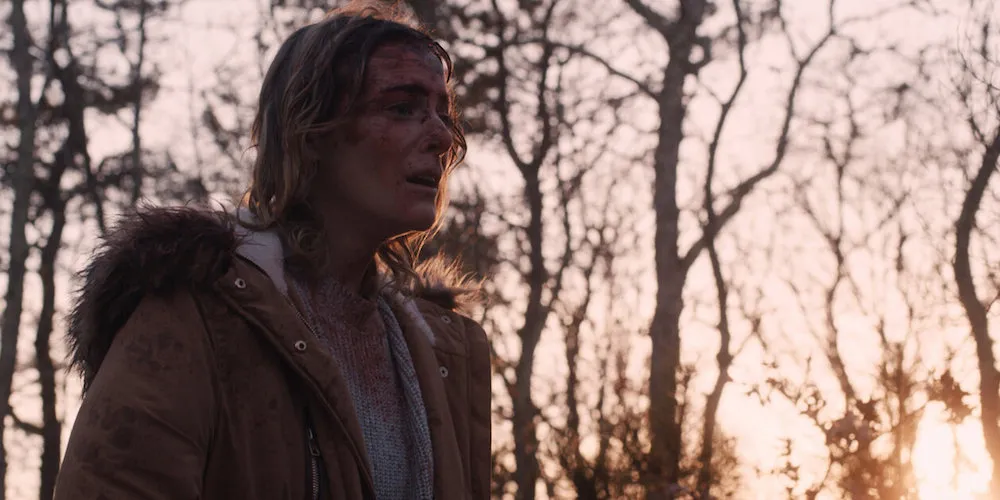 A white woman walking in the woods covered in blood.