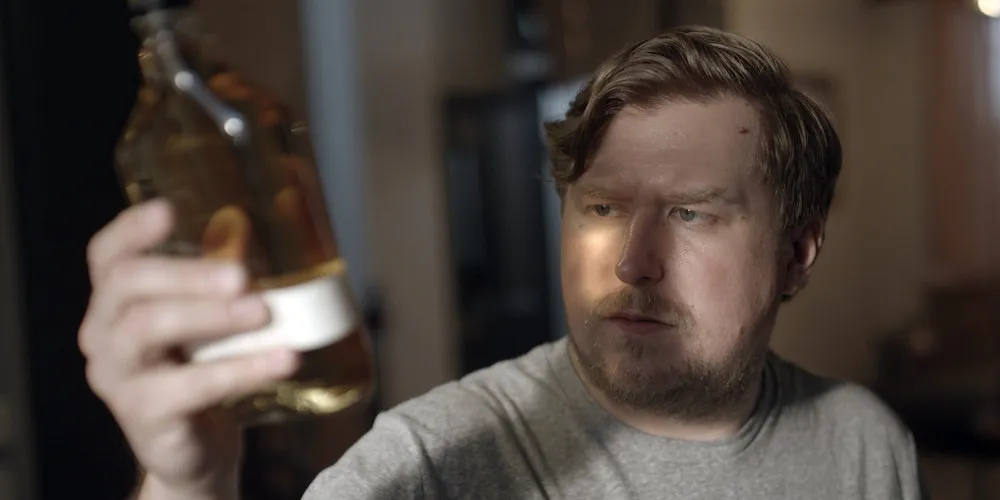 Still from a movie of a blond man holding up a bottle of liquor, looking puzzled. 