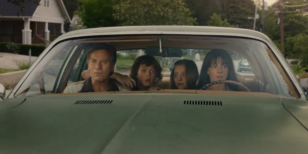 Film still from 'The End of Oak Street.' A family sits inside a green car driving through a suburban neighborhood. 