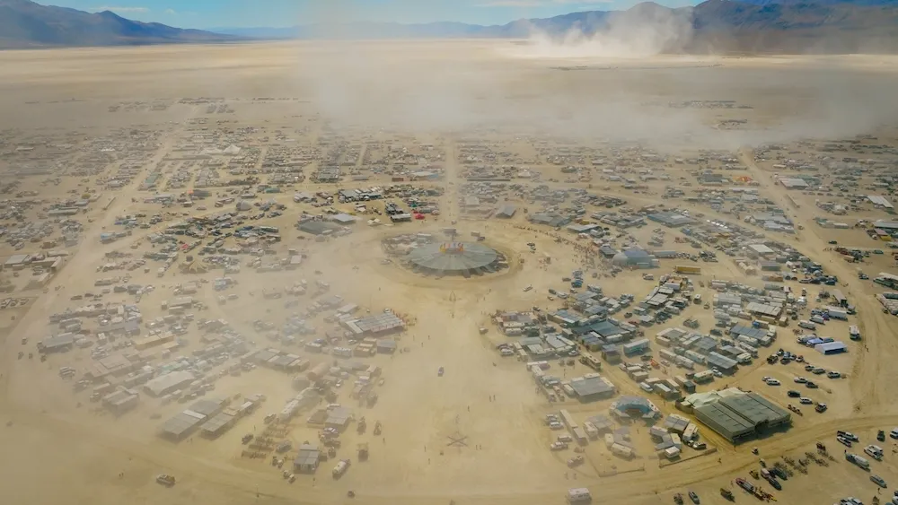 Aerial view of Black Rock City's circular grid of camps and dust clouds during Burning Man in the Nevada desert.