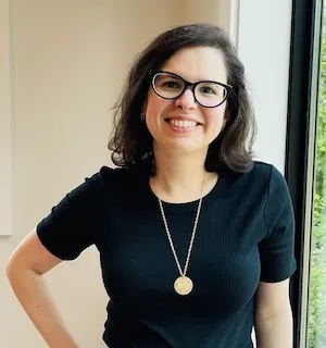 A white woman with brown hair and glasses smiling by a window, hand on hip.