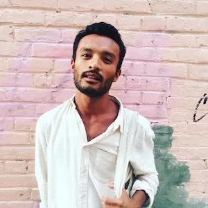 Man of color with facial hair in a white v neck shirt standing in front of pink and peach bricks. 