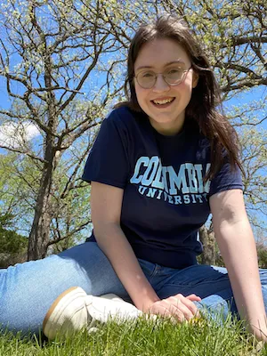 A girl with glasses and brown hair in a T-shirt sits on a lawn smiling.