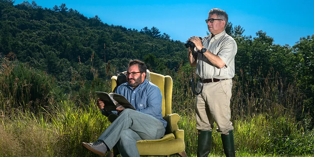 Two men outside, one on a wingback chair reading and the other looks like a bird watcher. 