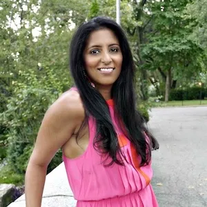 South Asian woman standing in front of bushes, smiling, wearing pink, long black hair