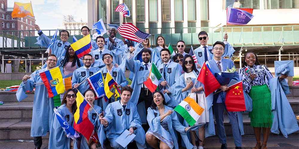 students in light blue cap and gowns for college graduation holding flags from different countries
