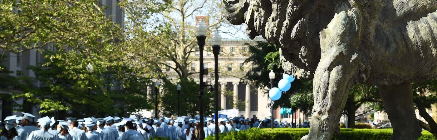 Statue of lion in the foreground, and a line of blue-clad graduates lining up in the background. 