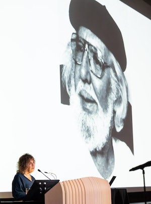 Dean Carol Becker stands at a podium with an image of a priest projected behind her