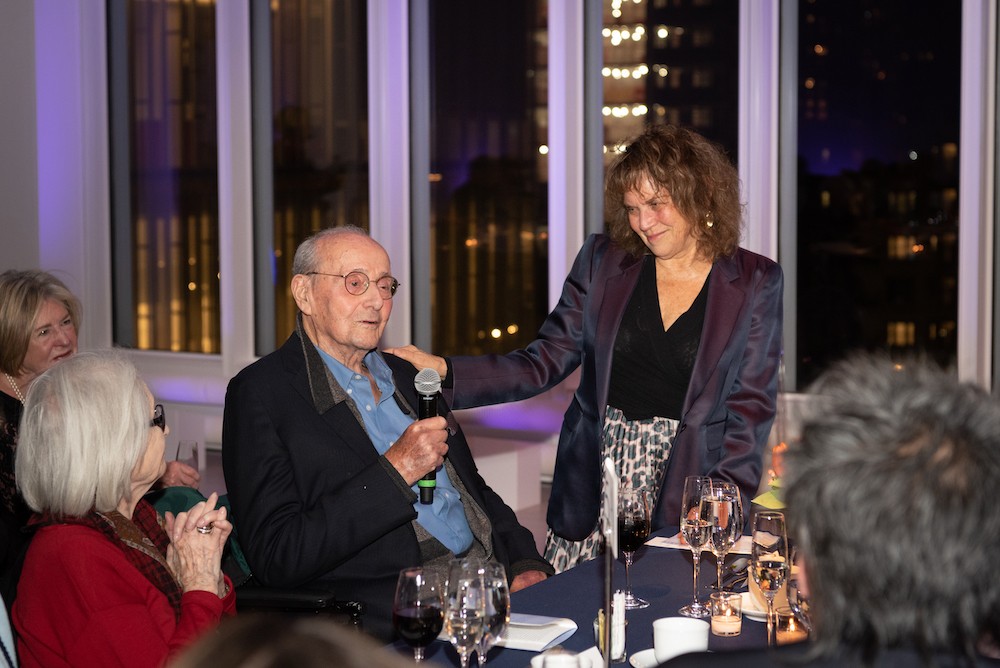 A seated man speaks into a microphone while a woman looks on. 