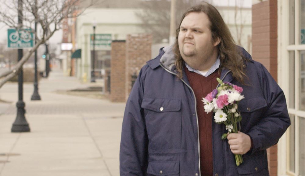 Still from 'Must Love Pie' Man with long hair holding flowers on sidewalk