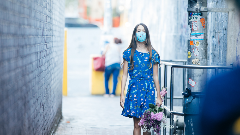 woman in blue dress with mask holding flowers in an alley 