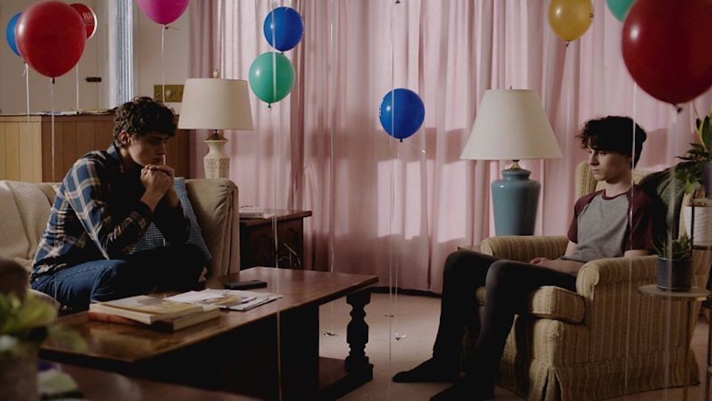 Still from Stay Awake - two young men sit solemnly in a living room filled with balloons. 