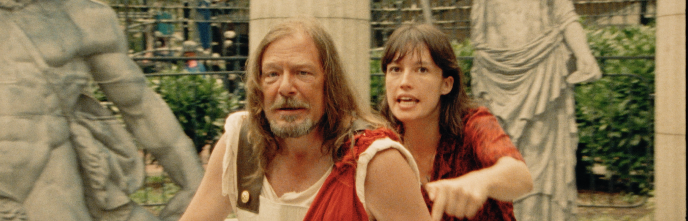 A Young woman stands behind and speaks to an older man wearing period ancient Roman costume. 
