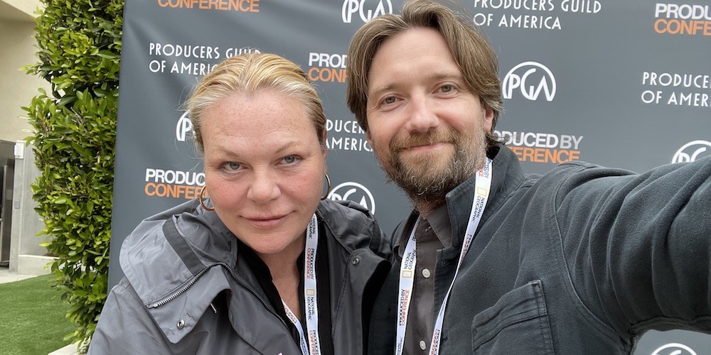 A woman and man stand in front of a step & repeat reading "Producers Guild of America"