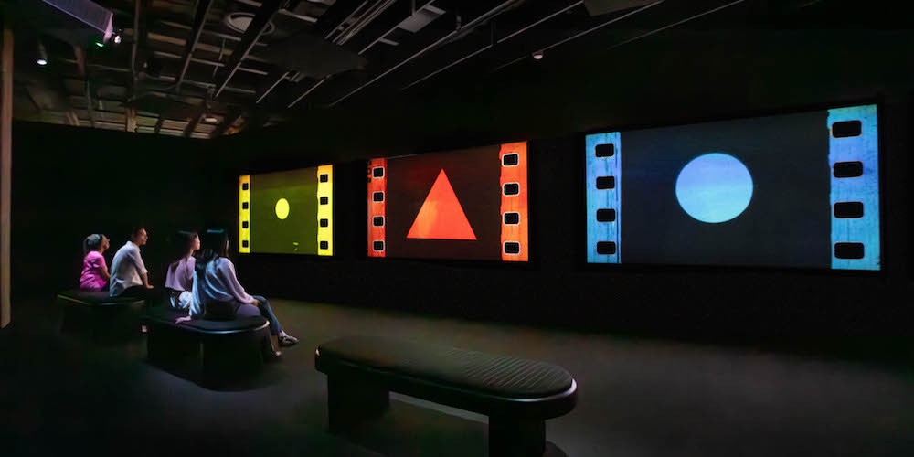 Viewers sitting on a bench in a dark gallery looking at three screens displaying a yellow circle, red triangle, and a blue dot. 