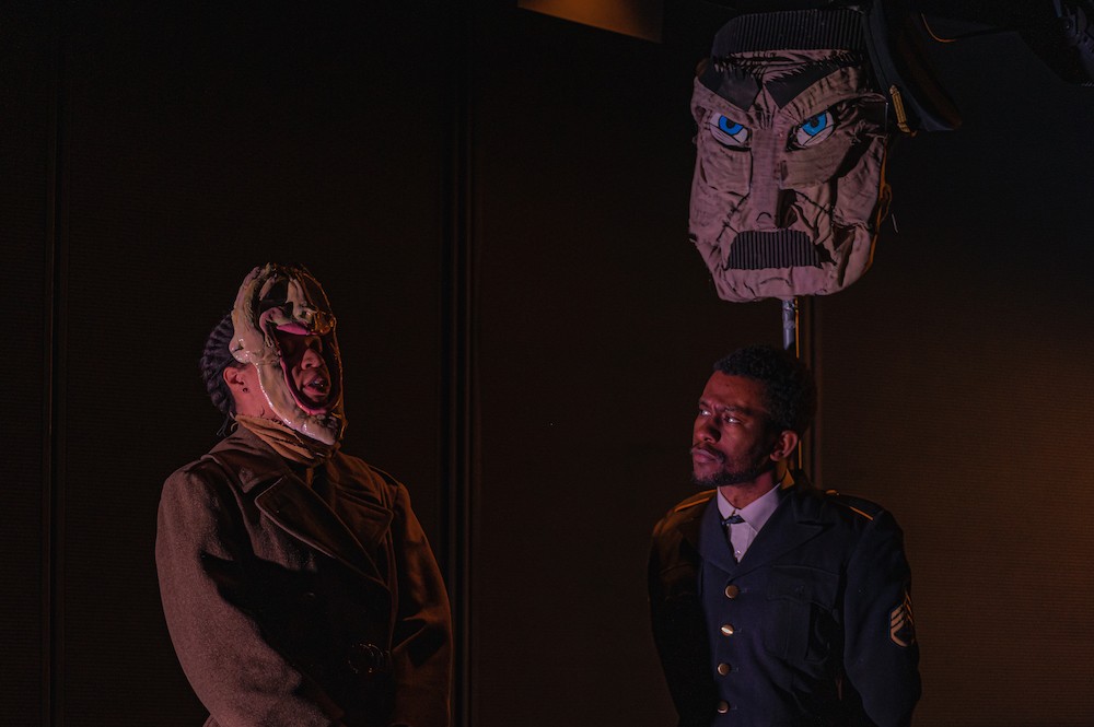 Two men stand in semi-darkness, wearing stylized masks. 