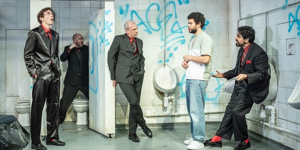 Several men stand in a stage made to look like a men's restroom. 