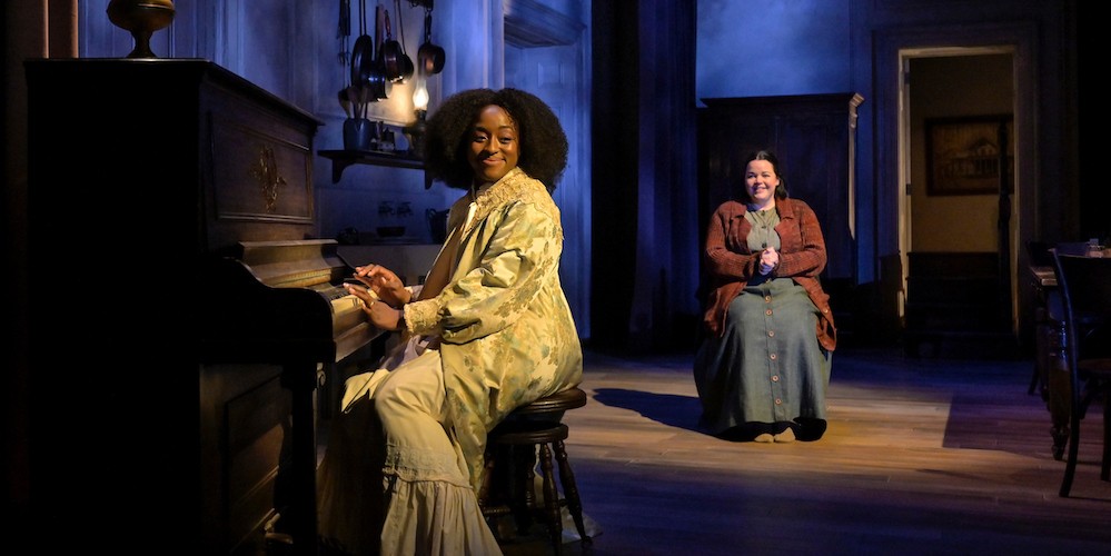 Two women in old-fashioned clothing sit on a stage made to look like a drawing room.