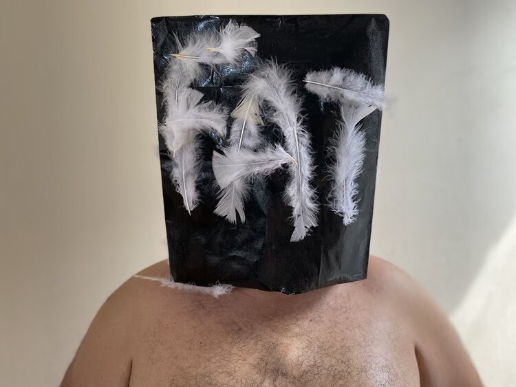 Shirtless person wearing a paper bag over their head with the word 'fat' spelled in white feathers
