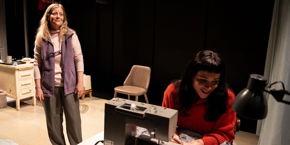 Two women on a stage - one works at a sewing machine and the other looks on affectionately. 