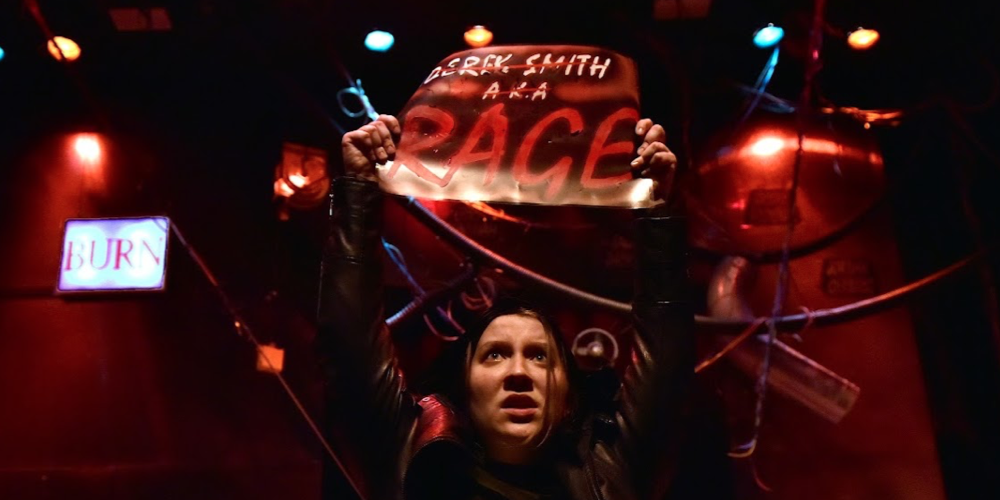 still image of character holding up a sign under red stage lights
