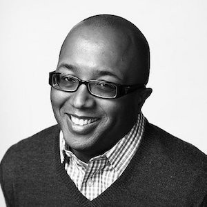 A Black man with black glasses and a v-neck sweater, smiling. 