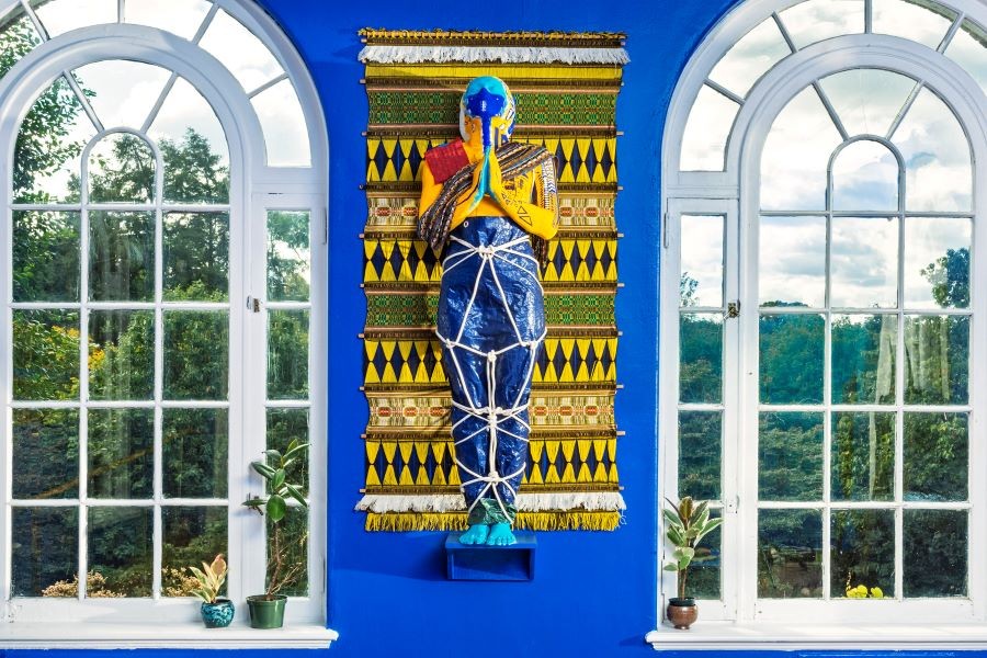 Origins of Devotions (Installation Detail), Dimensions Variable, Plaster, Blue Hurricane tarp as veil, Collected palm boots, Coconuts, Mamey seeds, Avocados from the Deering Estate, Tree snails collected by grandparents, Fragment of US-1, Palm tree leaves statue in front of yellow tapestry on blue wall flanked by windows