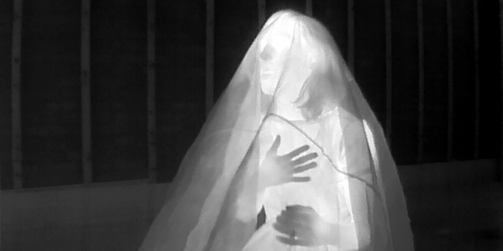 Still from video artwork by Asif Mian - a ghostly luminous figure, captured by thermal camera, sits under a shroud. 