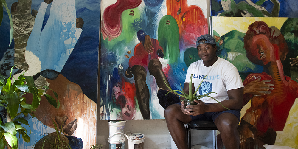 Khari Turner posing in front of his paintings holding a plant.