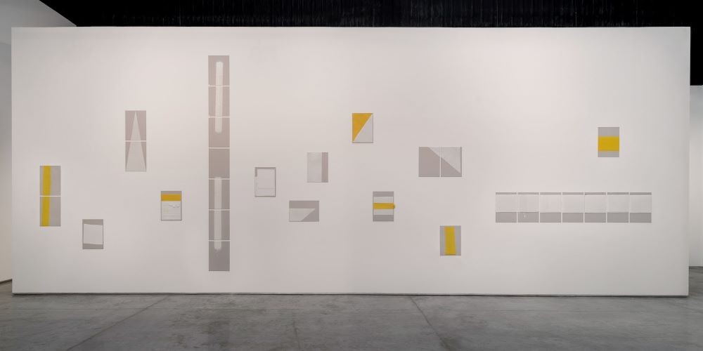 The art exhibit is mounted on a white wall. The art piece is composed of different canvasses with gray, white, and yellow.