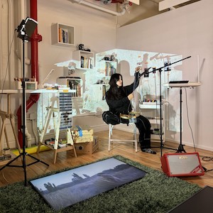 Woman sitting in a chair in an art studio surrounded by electronics and art supplies with a projection of an image on her. 