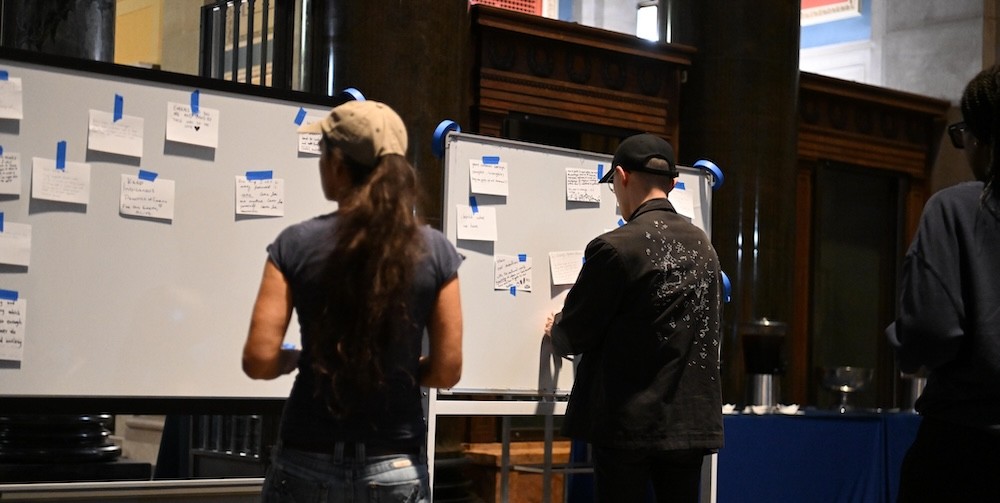 People stand in front of a cork board posting notes. 