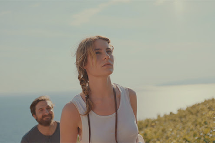 A blond woman walking through dunes with a man and body of water behind her.