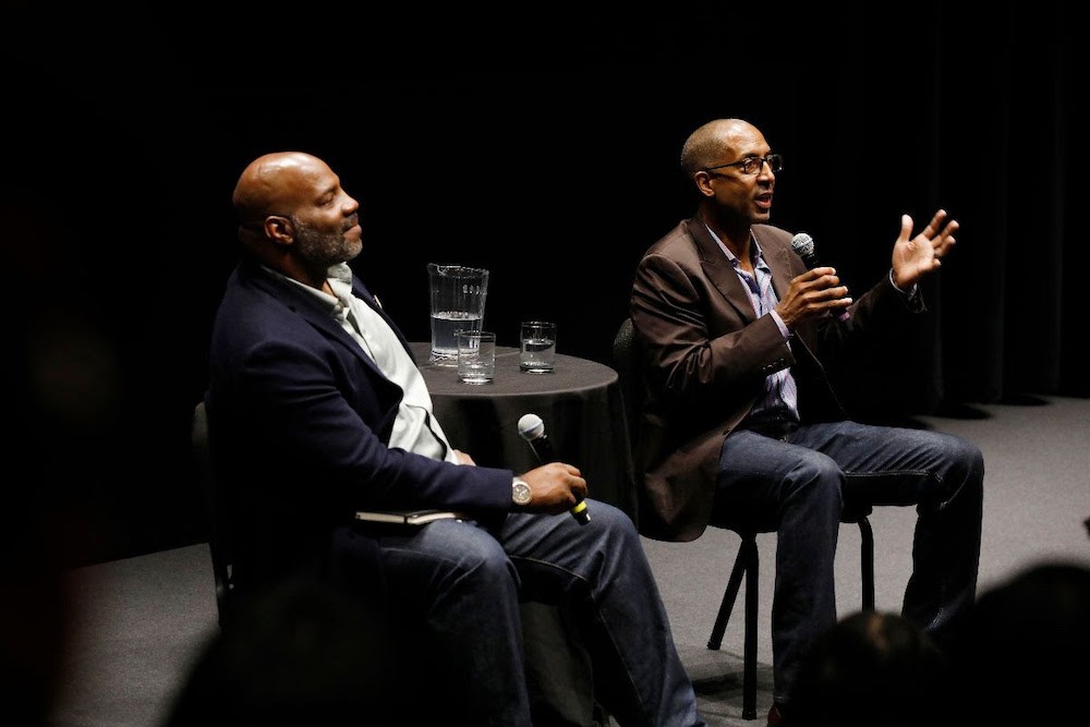 Two Black men speaking on stage. 