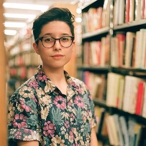 A light-skinned person with short hair and glasses in a library 