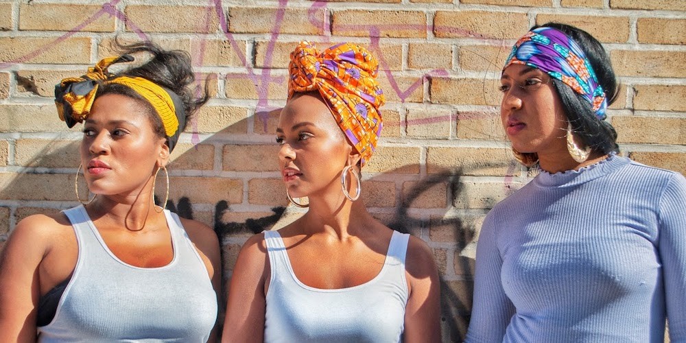 Three Black women in head wraps by a brick wall looking to the left.  