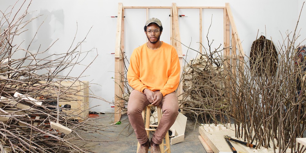 Black man in his studio wearing a hat and an orange sweatshirt surrounded by piles of branches.