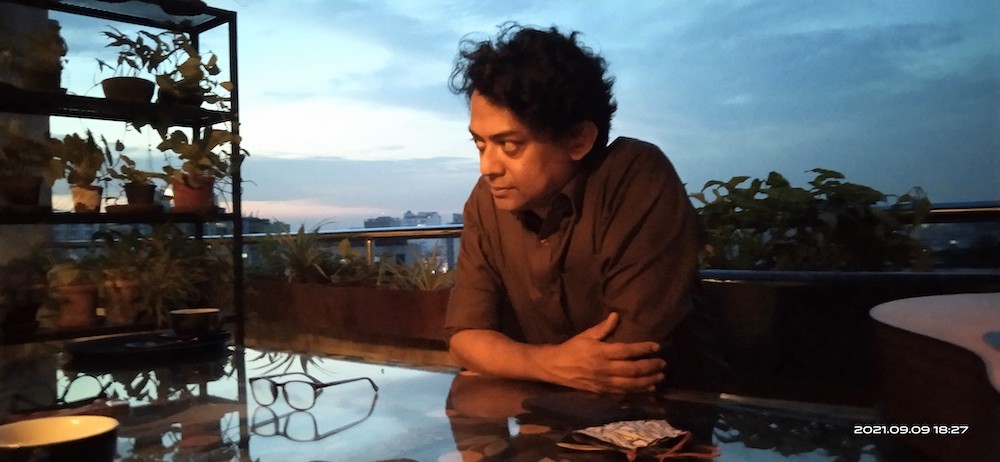 A South Asian man sitting on a patio, arms cross, looking in the distance.