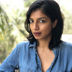 South Asian woman with collar-length hair and a denim shirt, purple lipstick, smiling