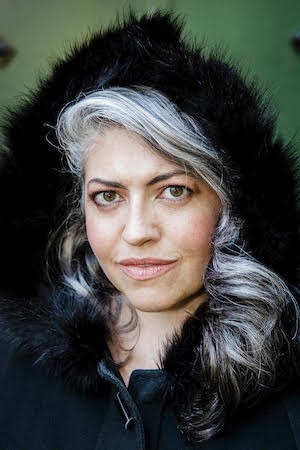 A light-skinned woman with wavy silver hair and an elegant black hood.